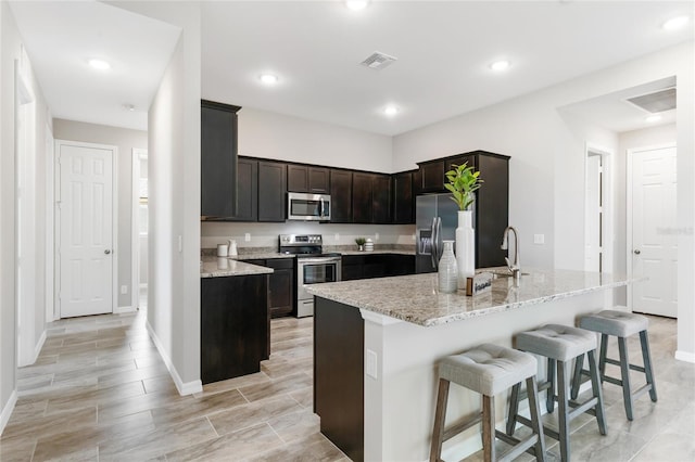 kitchen with a kitchen island with sink, sink, a kitchen bar, appliances with stainless steel finishes, and light stone counters