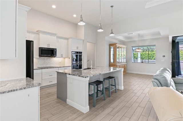 kitchen featuring black appliances, light wood-type flooring, white cabinetry, light stone counters, and a center island with sink