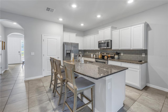 kitchen featuring decorative backsplash, white cabinets, a center island with sink, appliances with stainless steel finishes, and dark stone counters