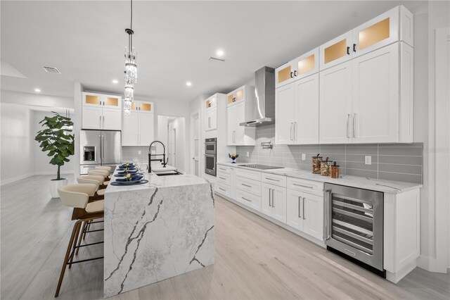 kitchen featuring beverage cooler, appliances with stainless steel finishes, white cabinetry, wall chimney range hood, and a kitchen island with sink