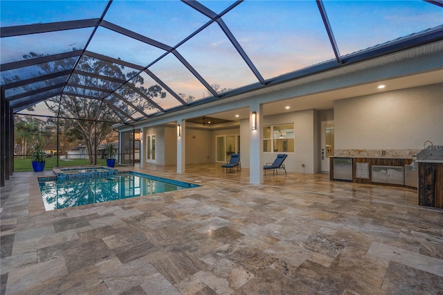 pool at dusk with glass enclosure, a patio area, an in ground hot tub, and an outdoor kitchen