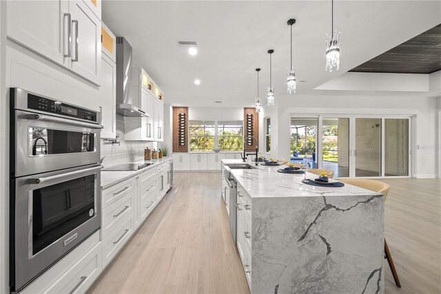 kitchen with light stone countertops, white cabinetry, a large island, stainless steel double oven, and wall chimney exhaust hood