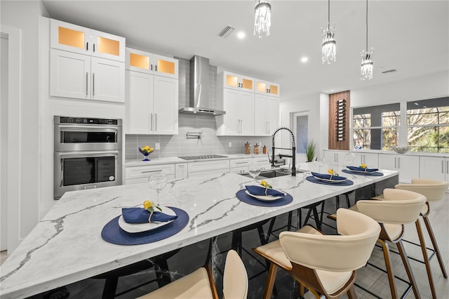 kitchen featuring double oven, wall chimney exhaust hood, sink, light stone countertops, and an island with sink