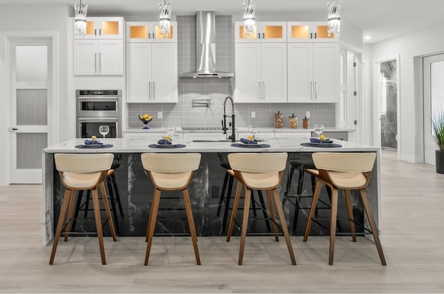 kitchen featuring white cabinets, stainless steel double oven, wall chimney exhaust hood, and a large island with sink