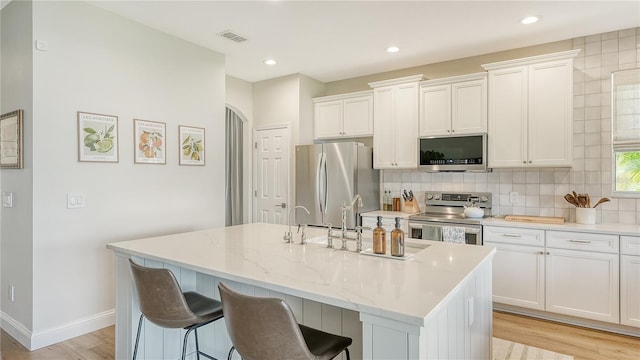 kitchen featuring appliances with stainless steel finishes, white cabinetry, a kitchen island with sink, light stone countertops, and light hardwood / wood-style flooring