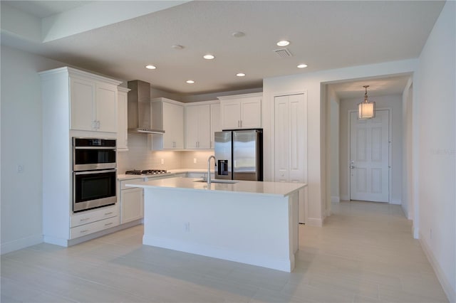 kitchen with white cabinets, light countertops, appliances with stainless steel finishes, wall chimney exhaust hood, and an island with sink