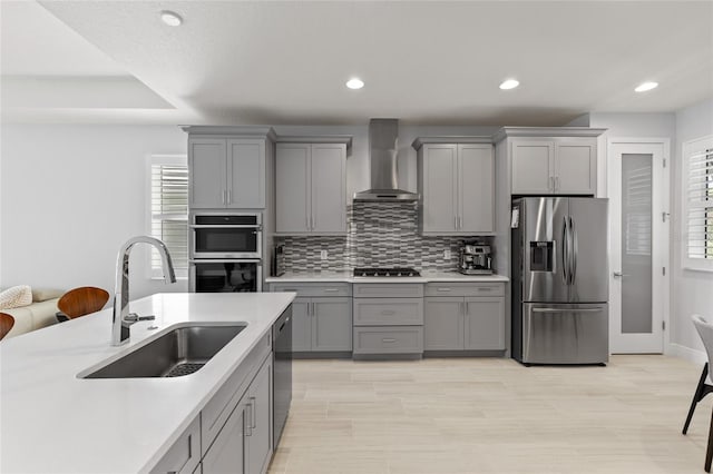 kitchen with wall chimney range hood, backsplash, appliances with stainless steel finishes, gray cabinets, and sink