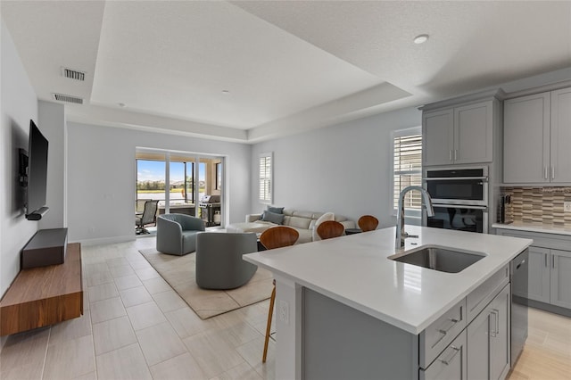 kitchen featuring gray cabinetry, appliances with stainless steel finishes, sink, an island with sink, and a tray ceiling