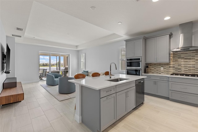 kitchen with a tray ceiling, wall chimney range hood, sink, and a kitchen island with sink