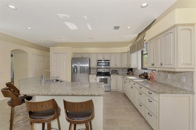 kitchen featuring an island with sink, backsplash, sink, light stone countertops, and appliances with stainless steel finishes