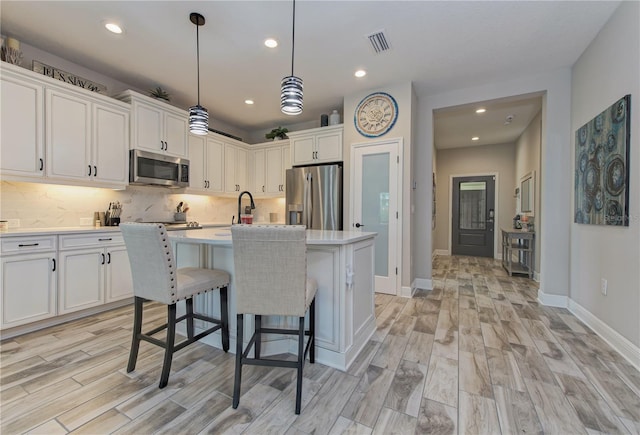 kitchen featuring white cabinets, hanging light fixtures, a center island with sink, appliances with stainless steel finishes, and light wood-type flooring