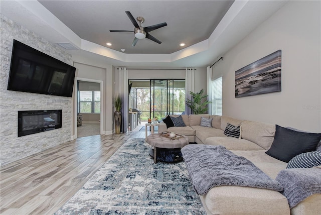 living room featuring ceiling fan, a stone fireplace, light wood-type flooring, and a raised ceiling