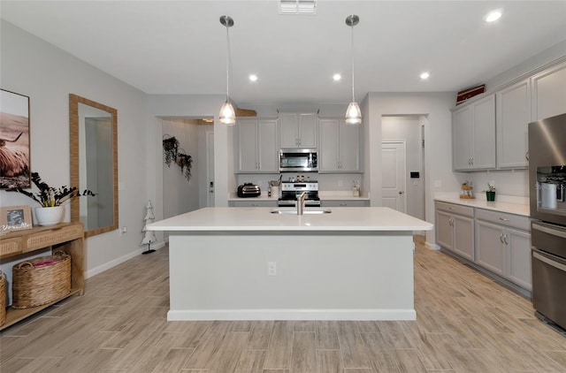 kitchen with decorative light fixtures, stainless steel appliances, light wood-type flooring, and an island with sink