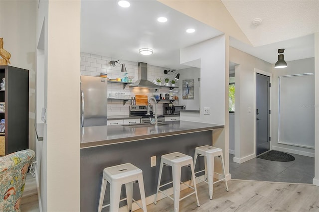 kitchen with wall chimney range hood, kitchen peninsula, appliances with stainless steel finishes, a kitchen bar, and light wood-type flooring