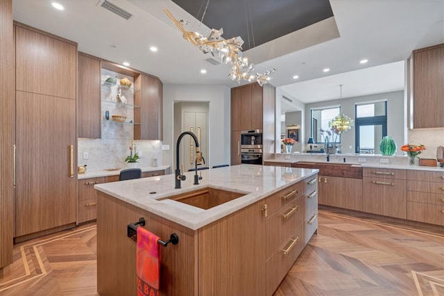 kitchen with sink, backsplash, light parquet floors, light brown cabinetry, and a center island with sink