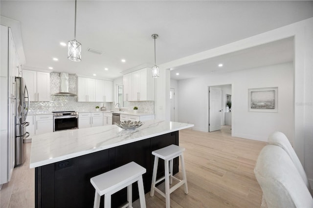 kitchen with white cabinetry, wall chimney range hood, appliances with stainless steel finishes, decorative light fixtures, and light wood-type flooring