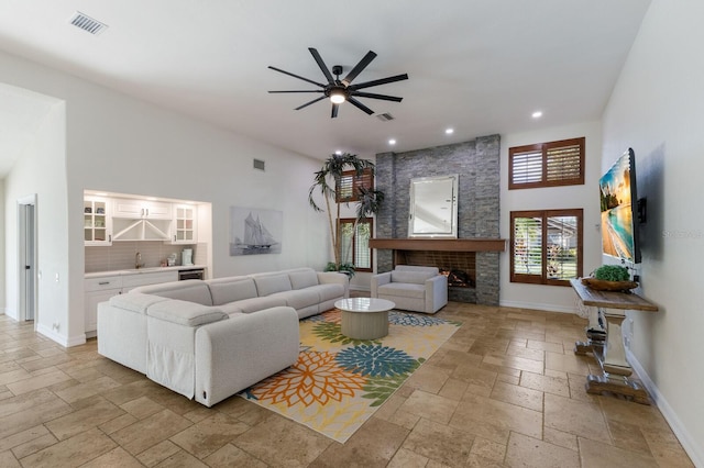 living room with ceiling fan, a high ceiling, a stone fireplace, and indoor wet bar