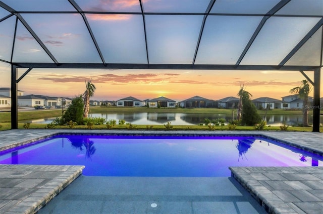 pool at dusk with glass enclosure, a patio area, and a water view