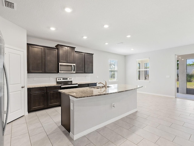 kitchen with a kitchen island with sink, dark brown cabinets, sink, appliances with stainless steel finishes, and light stone counters