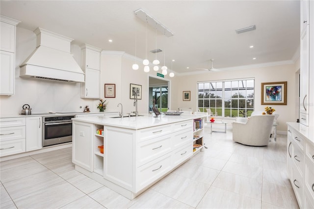 kitchen with white cabinetry, an island with sink, custom range hood, decorative light fixtures, and stainless steel oven