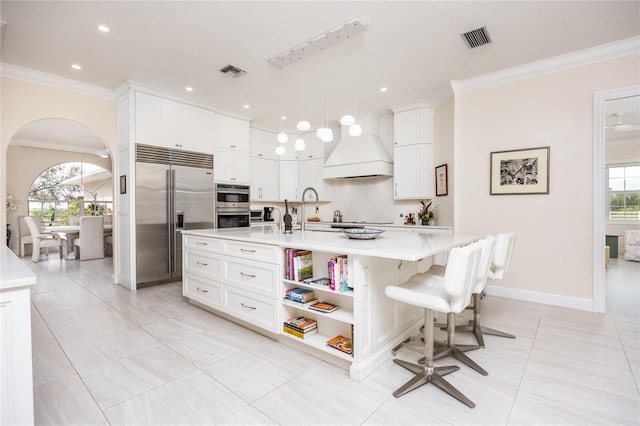 kitchen with hanging light fixtures, stainless steel appliances, an island with sink, white cabinets, and custom exhaust hood