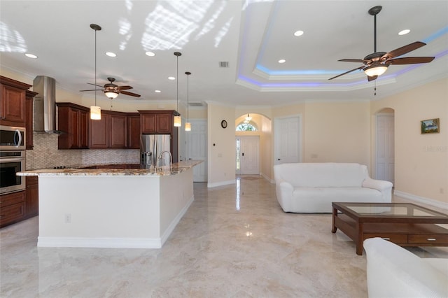 kitchen with backsplash, stainless steel appliances, a kitchen island with sink, wall chimney range hood, and decorative light fixtures