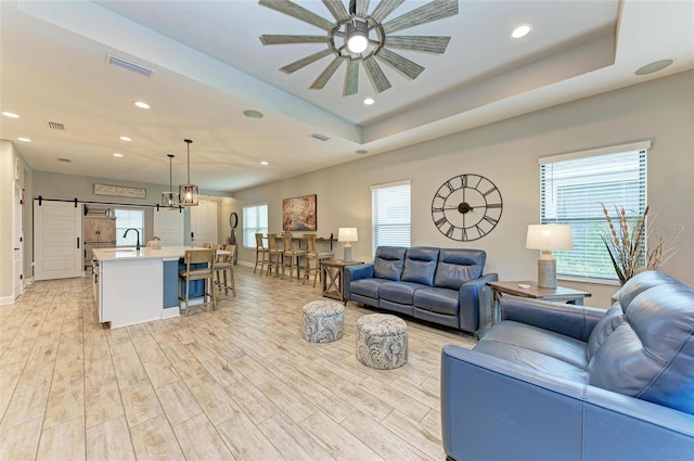 living room featuring a barn door, sink, ceiling fan, a tray ceiling, and light wood-type flooring