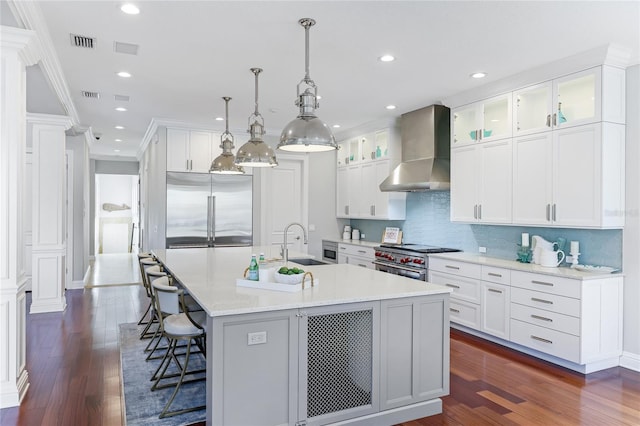 kitchen featuring high end appliances, hanging light fixtures, a kitchen island with sink, white cabinets, and wall chimney exhaust hood