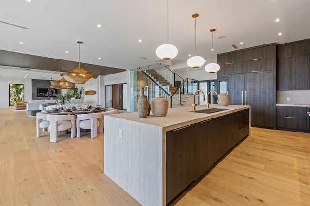 kitchen with sink, dark brown cabinets, pendant lighting, light wood-type flooring, and a large island with sink