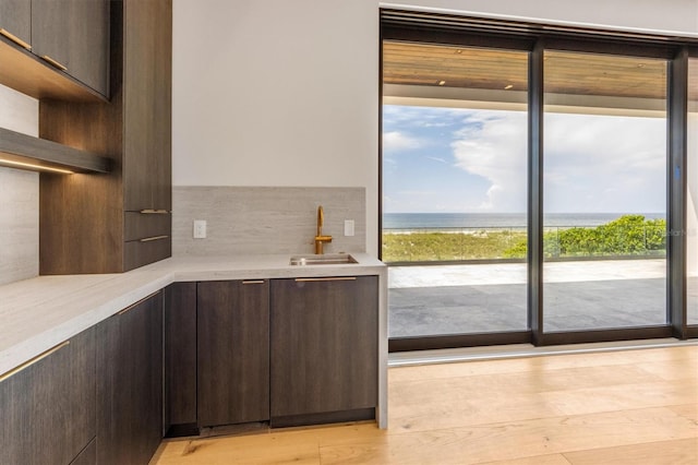 bar with backsplash, light wood-type flooring, dark brown cabinets, sink, and a water view