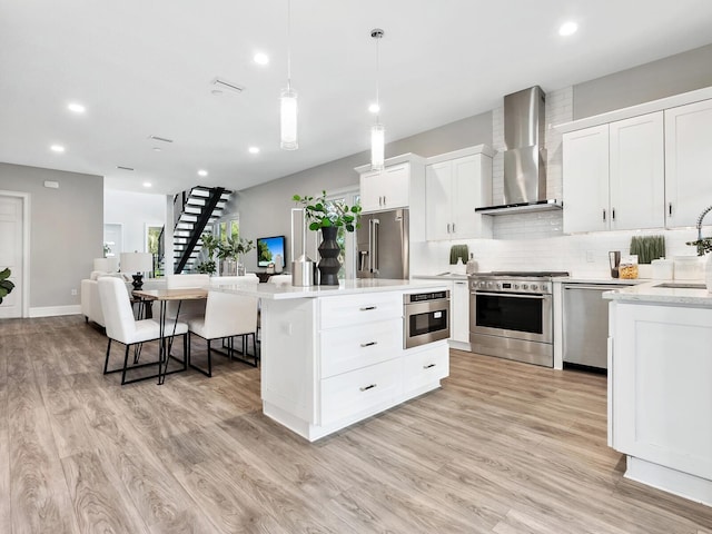 kitchen with wall chimney exhaust hood, high quality appliances, pendant lighting, light wood-type flooring, and white cabinets