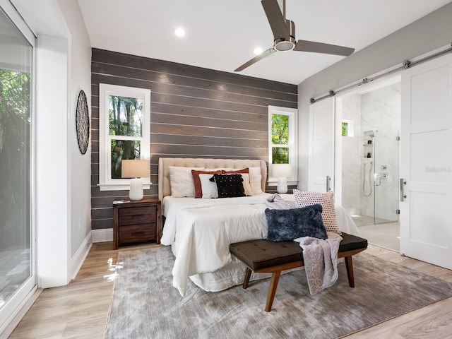 bedroom featuring connected bathroom, wooden walls, a barn door, light hardwood / wood-style floors, and ceiling fan