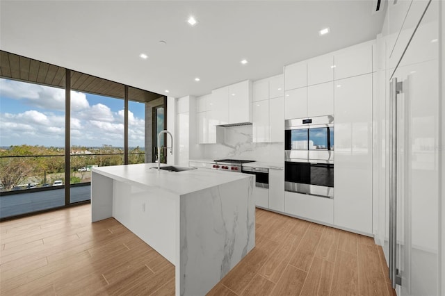 kitchen with white cabinets, sink, a kitchen island with sink, a wall of windows, and appliances with stainless steel finishes