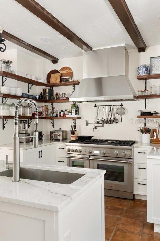 kitchen featuring wall chimney exhaust hood, beamed ceiling, light stone countertops, range with two ovens, and white cabinets