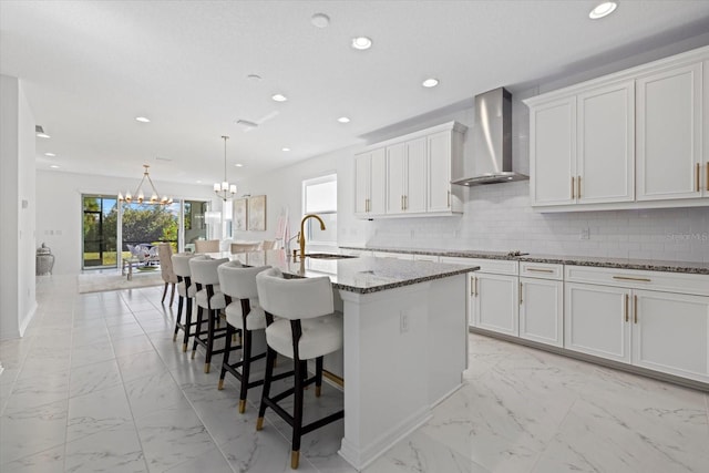 kitchen featuring white cabinets, wall chimney exhaust hood, sink, stone counters, and an island with sink