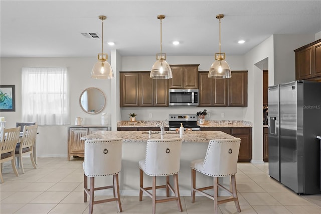 kitchen featuring a kitchen island with sink, light stone counters, decorative light fixtures, and appliances with stainless steel finishes