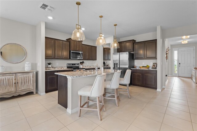 kitchen with pendant lighting, stainless steel appliances, visible vents, dark brown cabinets, and an island with sink