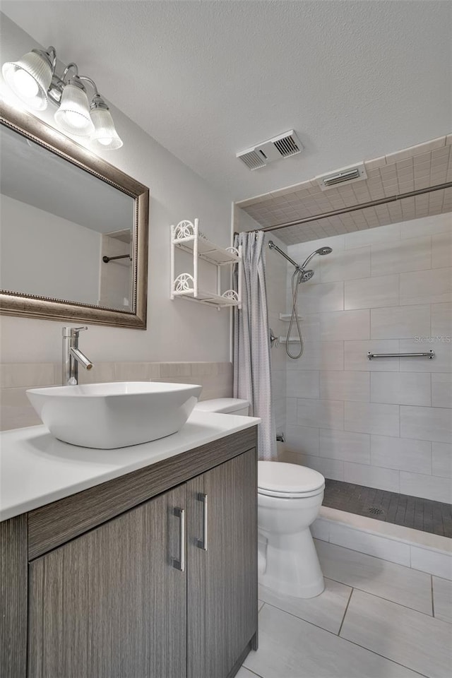 full bath with toilet, tiled shower, vanity, a textured ceiling, and visible vents