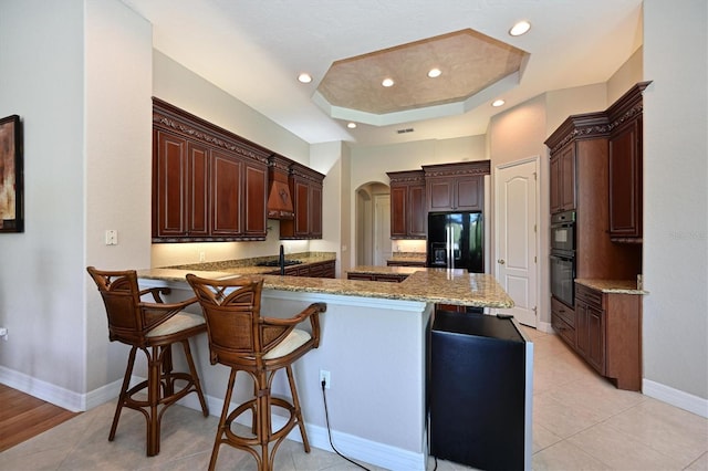 kitchen with black appliances, light wood-type flooring, a tray ceiling, kitchen peninsula, and a breakfast bar area