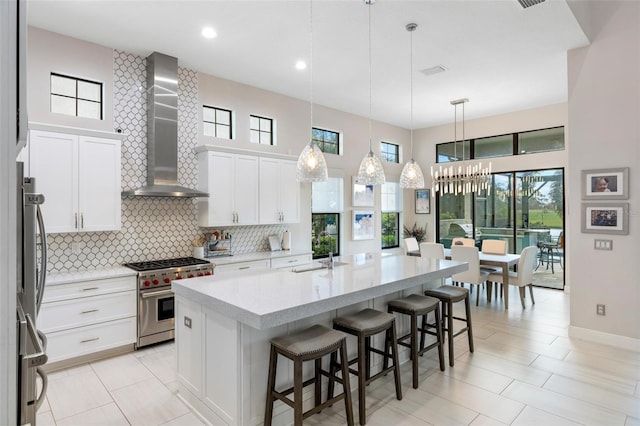 kitchen featuring wall chimney range hood, pendant lighting, a kitchen island with sink, white cabinets, and appliances with stainless steel finishes