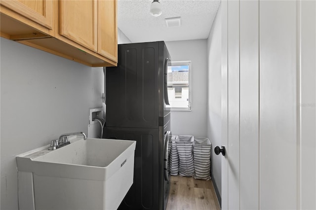 laundry area featuring a textured ceiling, stacked washer and dryer, cabinets, sink, and light hardwood / wood-style floors