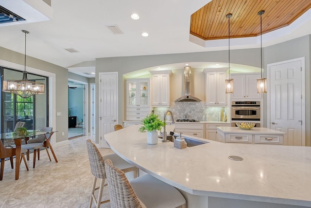 kitchen with a center island with sink, white cabinetry, tasteful backsplash, wall chimney range hood, and pendant lighting