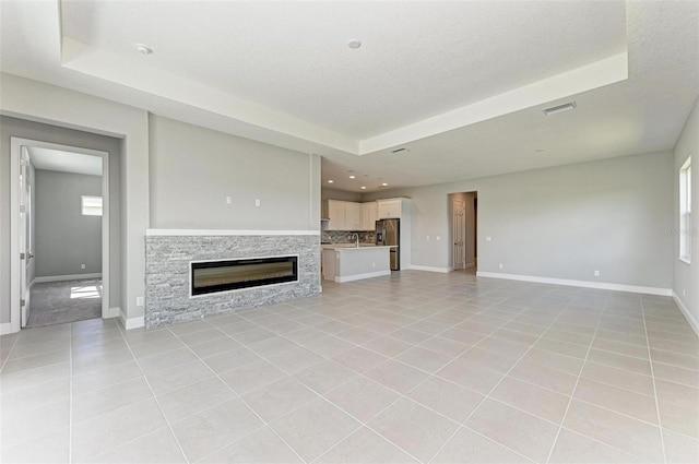 unfurnished living room with a stone fireplace, a textured ceiling, light tile patterned flooring, and a tray ceiling