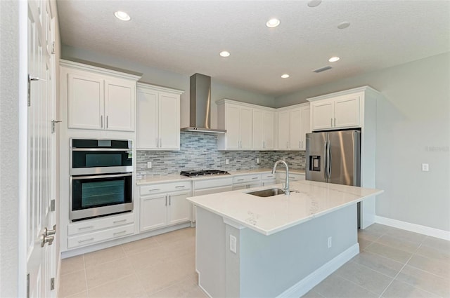 kitchen with sink, appliances with stainless steel finishes, wall chimney exhaust hood, an island with sink, and white cabinets