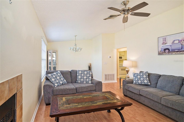 living room with light hardwood / wood-style floors, a tiled fireplace, ceiling fan with notable chandelier, a textured ceiling, and vaulted ceiling