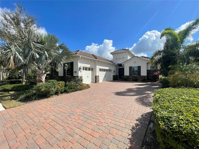 mediterranean / spanish home featuring decorative driveway, a tile roof, stucco siding, a garage, and stone siding