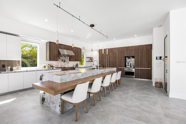 kitchen with decorative backsplash, wall chimney exhaust hood, stainless steel oven, pendant lighting, and white cabinets