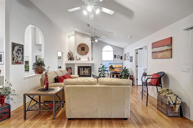 living room featuring light wood-type flooring, a textured ceiling, vaulted ceiling, a tile fireplace, and ceiling fan