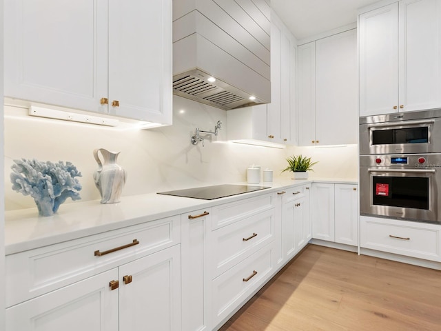 kitchen with black electric stovetop, light wood-type flooring, custom range hood, double oven, and white cabinetry