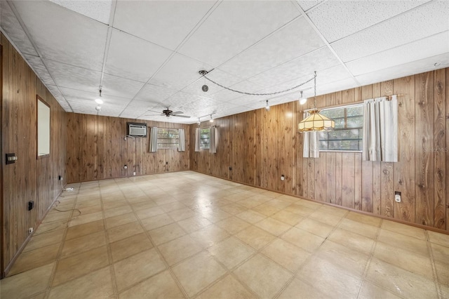 basement with a wall unit AC, a paneled ceiling, wooden walls, and ceiling fan